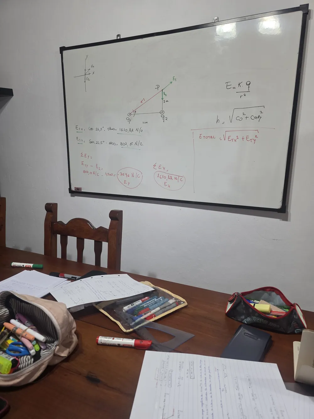 Pizarra con el desarrollo de un ejercicio de física junto a una mesa de estudio con apuntes y cartucheras.