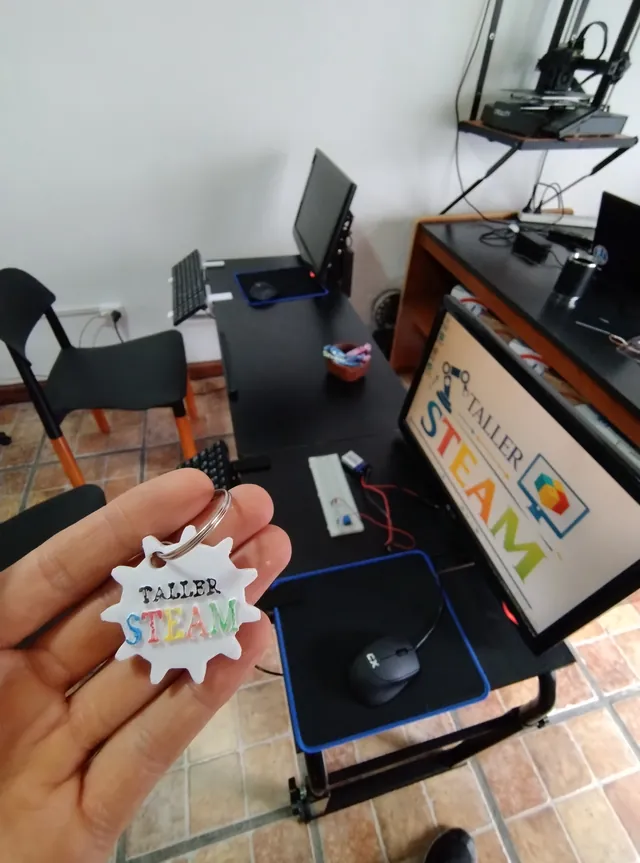 Un llavero impreso en 3D con el logo del taller, sostenido frente al aula de tecnología con sus computadoras y la impresora 3D.